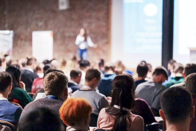 Une femme à cheveux longs fait une conférence dans une salle avec beaucoup de monde