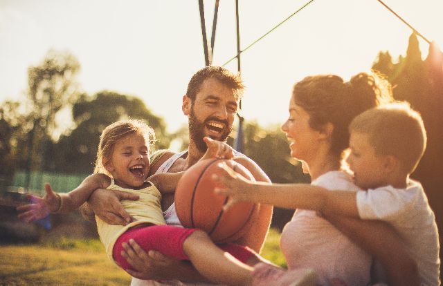 Famille qui joue avec un ballon de basket, tout le monde participe et s’amuse. La fille est dans les bras de son père.