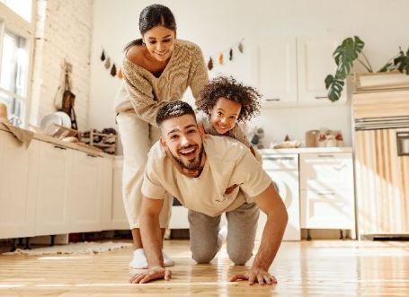 parents jouent par terre avec leur enfant dans la cuisine. L’enfant monte sur le dos de son père. Ils sont contents